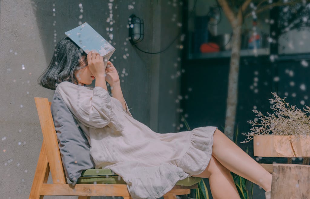 A young woman covers her face with a book while sitting outdoors on a rainy day, enjoying a peaceful moment.