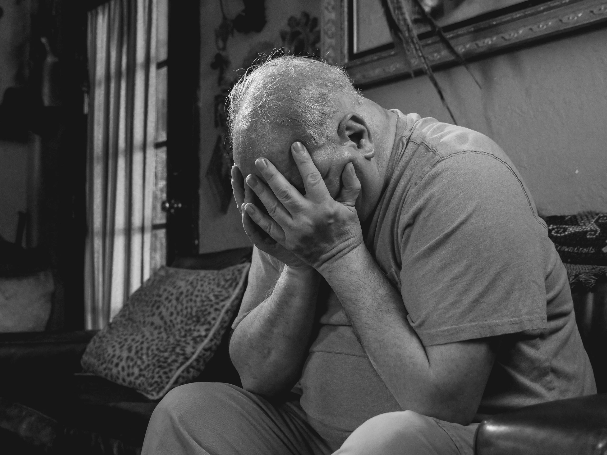 Black and white portrait of a senior man sitting indoors, expressing deep emotion and distress.