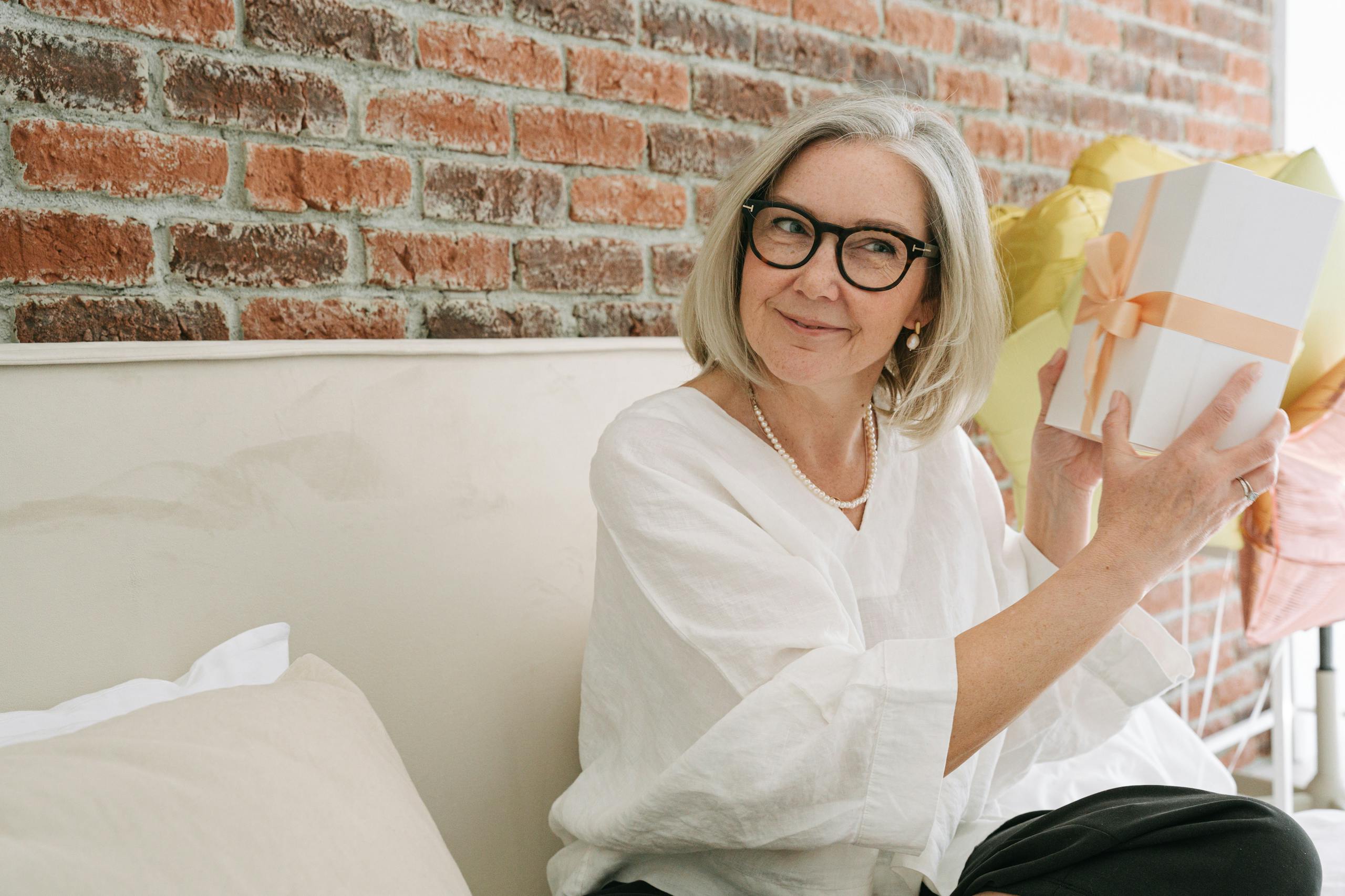 Senior woman in white blouse happily holding a gift box indoors in front of a brick wall.
