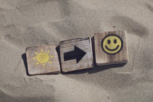Wooden blocks with sun, arrow, and smiley emoticons on sand, evoking summer vibes.