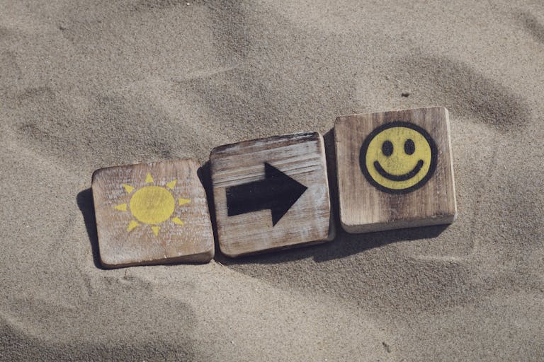 Wooden blocks with sun, arrow, and smiley emoticons on sand, evoking summer vibes.