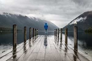Person in blue jacket stands on a rainy dock overlooking serene lake and mountains.