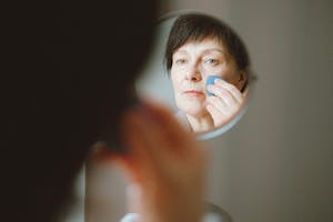 Elderly woman looking at herself in the mirror while applying makeup sponge.