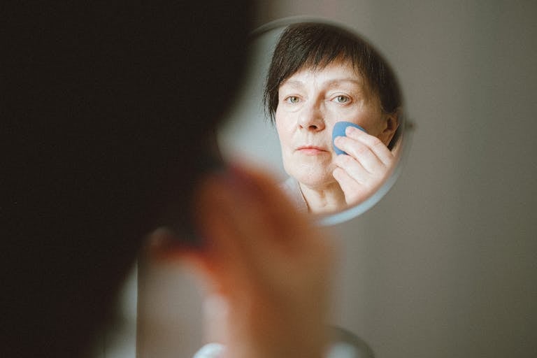 Elderly woman looking at herself in the mirror while applying makeup sponge.