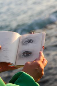 Close-up of an artist's hand holding a sketchbook with eye drawings by the ocean during sunset.