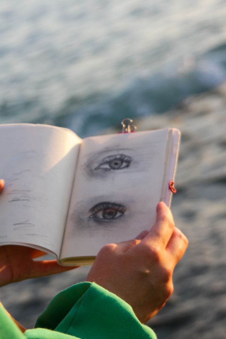 Close-up of an artist's hand holding a sketchbook with eye drawings by the ocean during sunset.