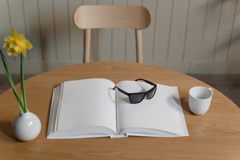 Open Braille book with sunglasses and daffodil on wooden table indoors.